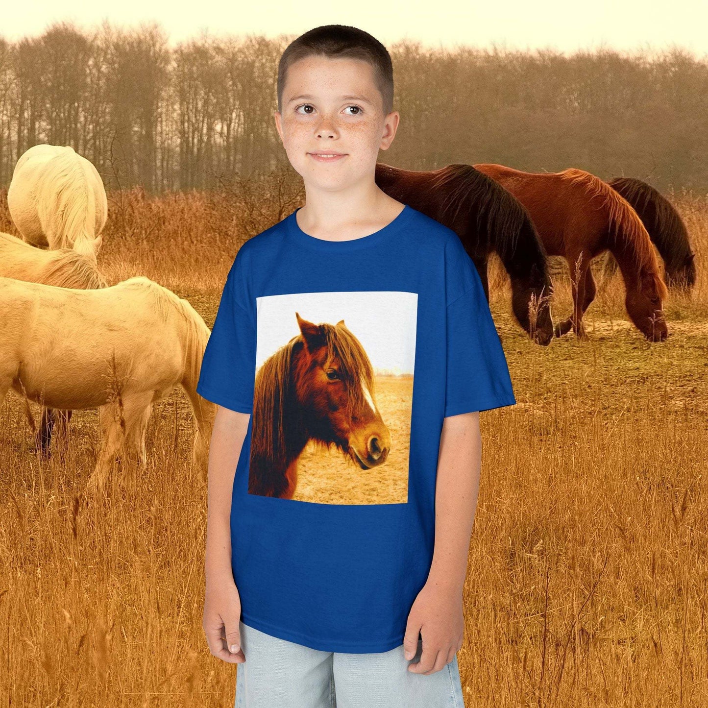 Boy wearing a blue kids tee with a horse print, standing in a field with horses in the background.