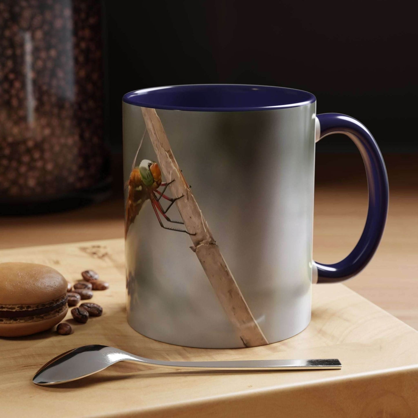 White ceramic mug with a dragonfly design, blue interior, on a wooden surface with coffee beans and a macaron.
