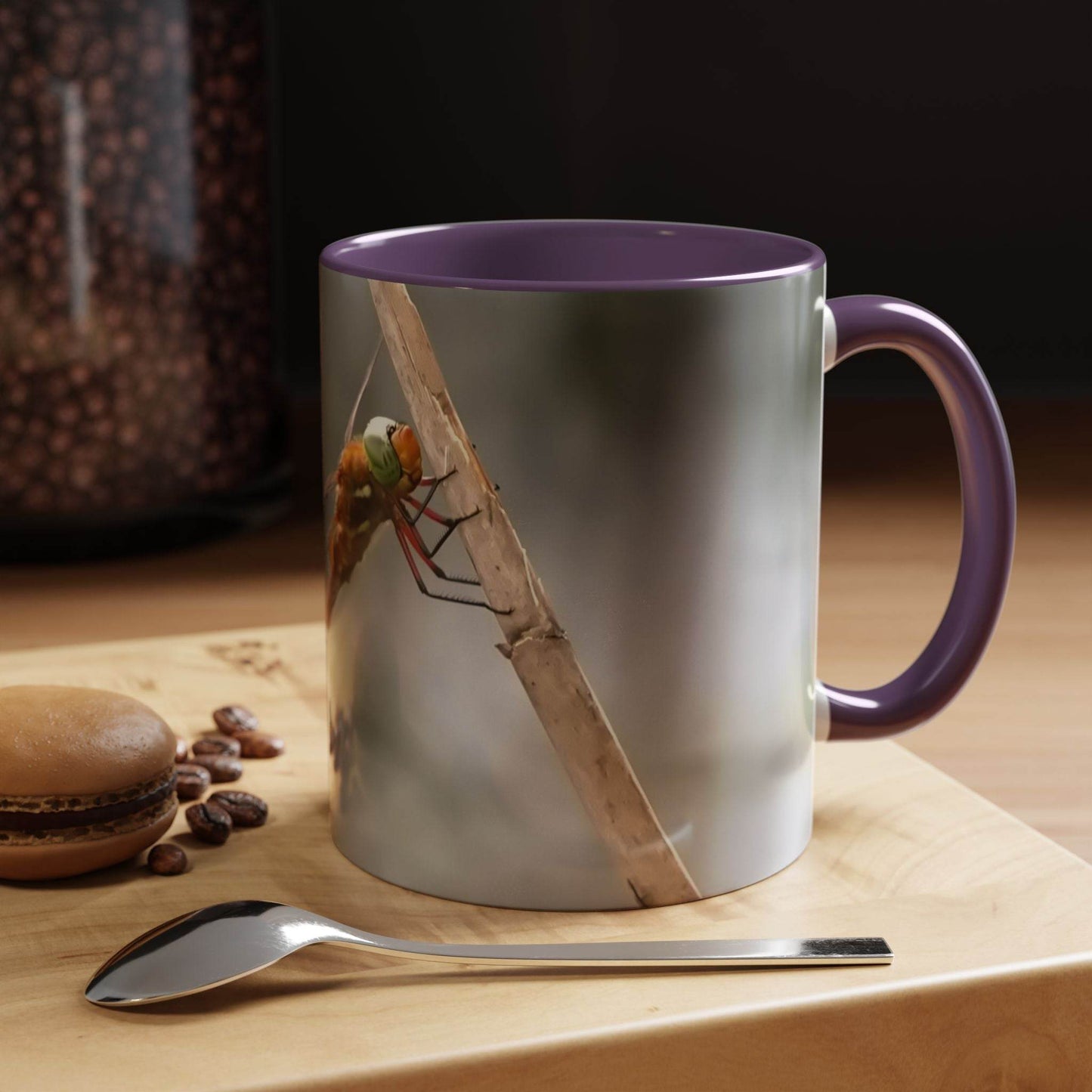 White ceramic mug with a dragonfly design, featuring a purple handle and glossy finish, placed on a wooden table with a spoon and coffee beans.